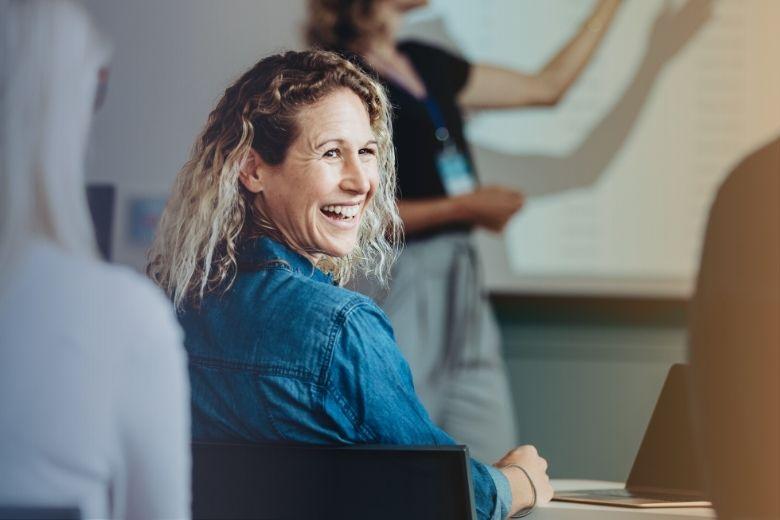 A woman in her 30s with blond hair smiles widely over her shoulder in a classroom