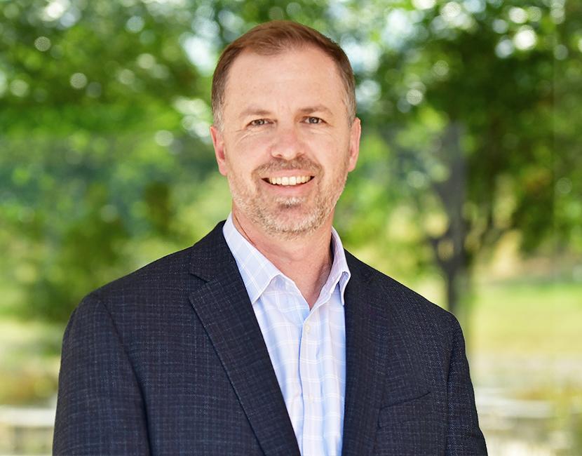 A professional headshot of a smiling man in a suit standing outdoors in front of a lushly wooded area. The collar of his button-up shirt is open, and he appears relaxed.
