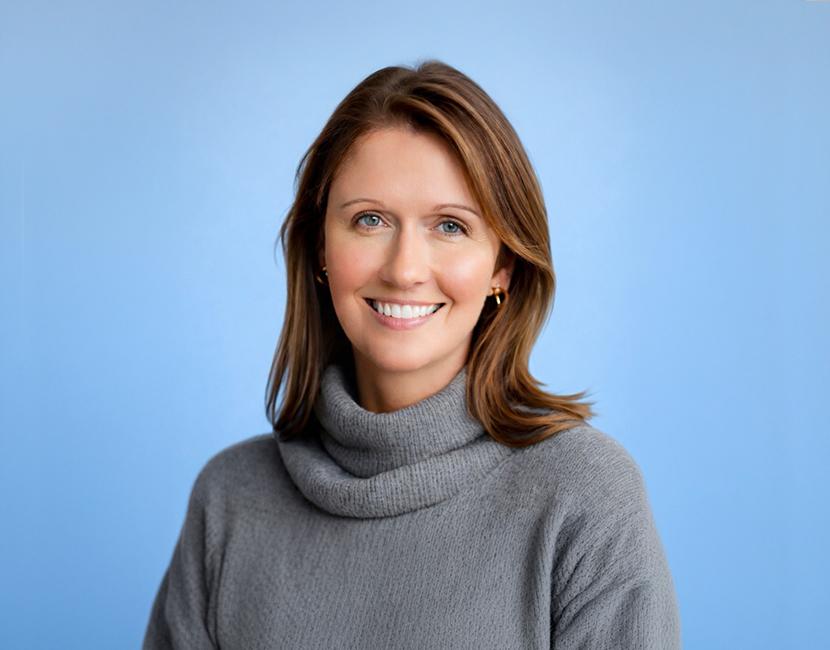 A professional headshot of a woman with mid length wavy brown hair in a black turtleneck in front of a marbled backdrop.