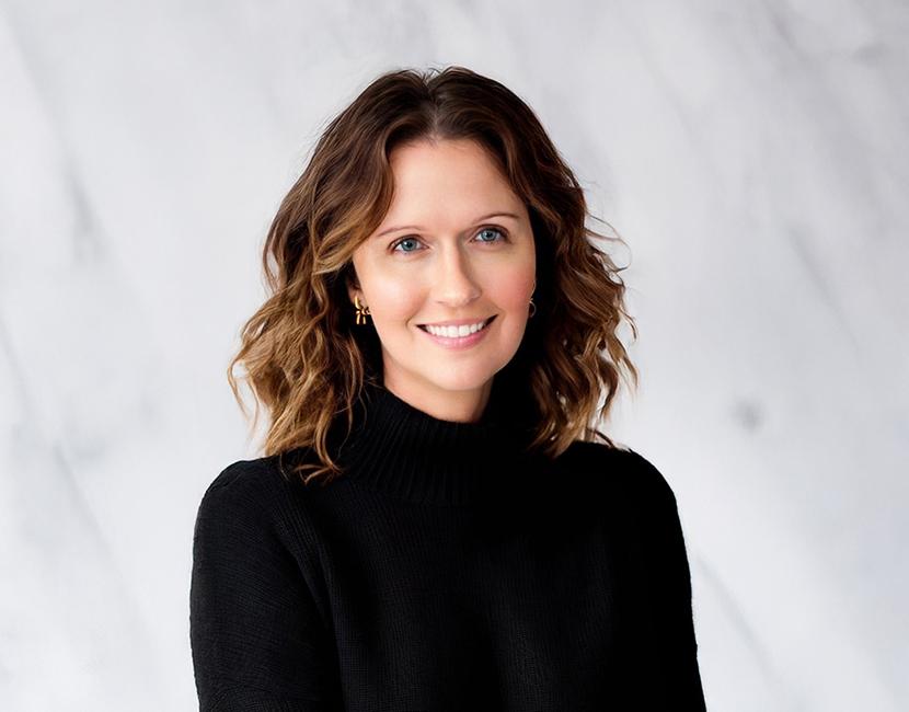 A professional headshot of a woman with mid length wavy brown hair in a black turtleneck in front of a marbled backdrop.
